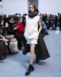 Runway model in a sleeveless white ribbed knit mini dress with a large dark hooded cape, walking the catwalk as the seated audience watches.
