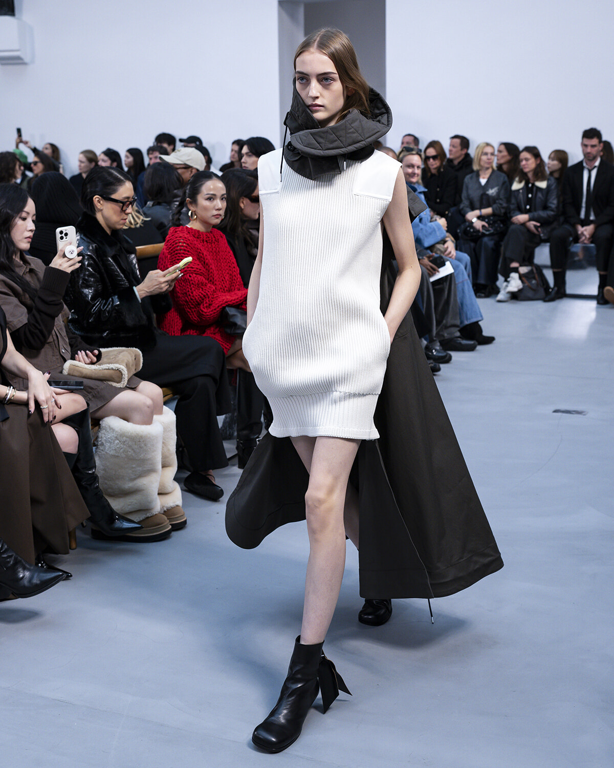 Runway model in a sleeveless white ribbed knit mini dress with a large dark hooded cape, walking the catwalk as the seated audience watches.