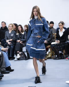 Model walks the runway in a patchwork denim ensemble with frayed edges and white stripe accents, paired with black fringe shoes.