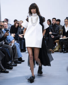 Model walks a white shift dress with a sheer black cape, layered pearls, and bold jewelry on a runway. Audience watches from the sides.