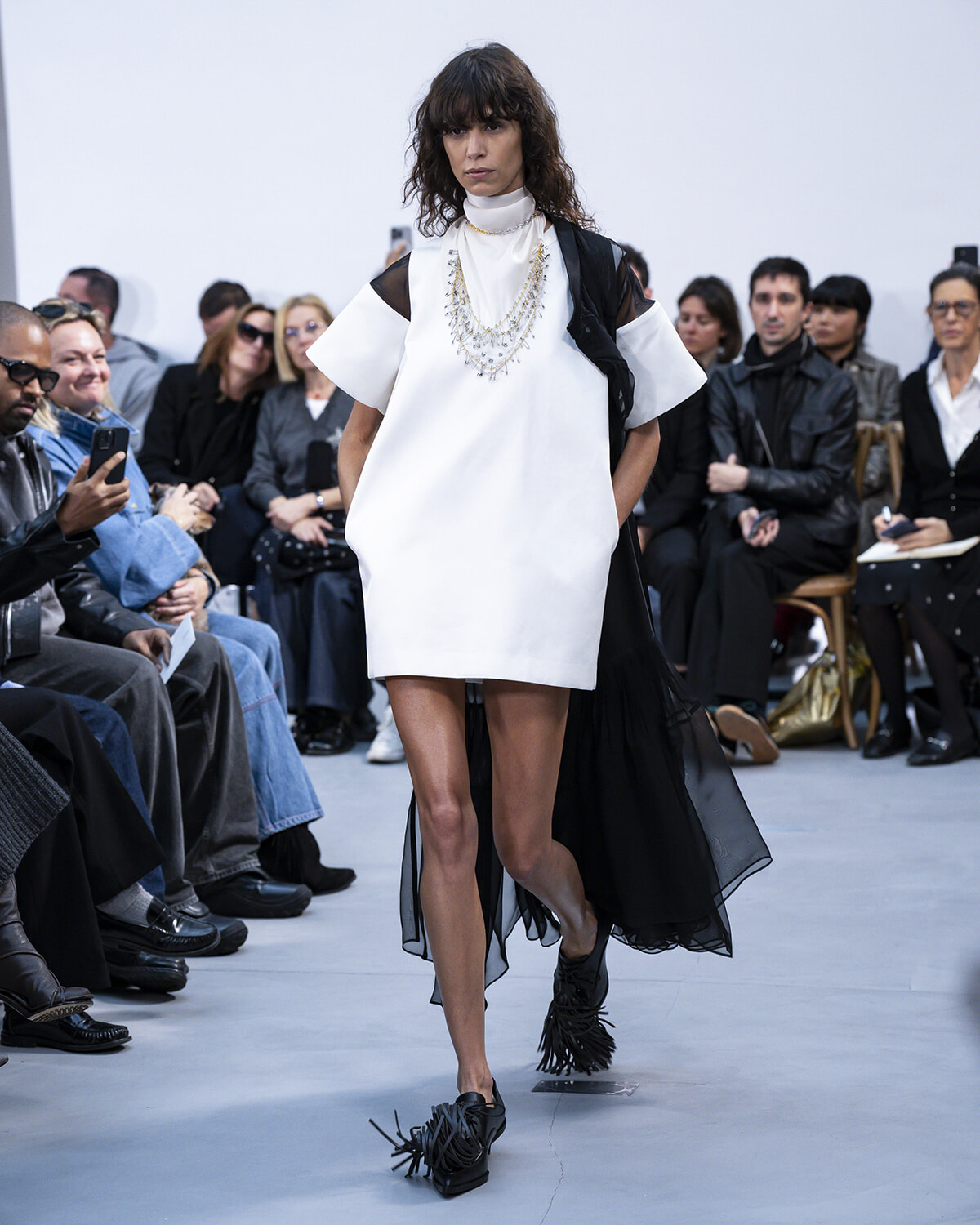 Model walks a white shift dress with a sheer black cape, layered pearls, and bold jewelry on a runway. Audience watches from the sides.