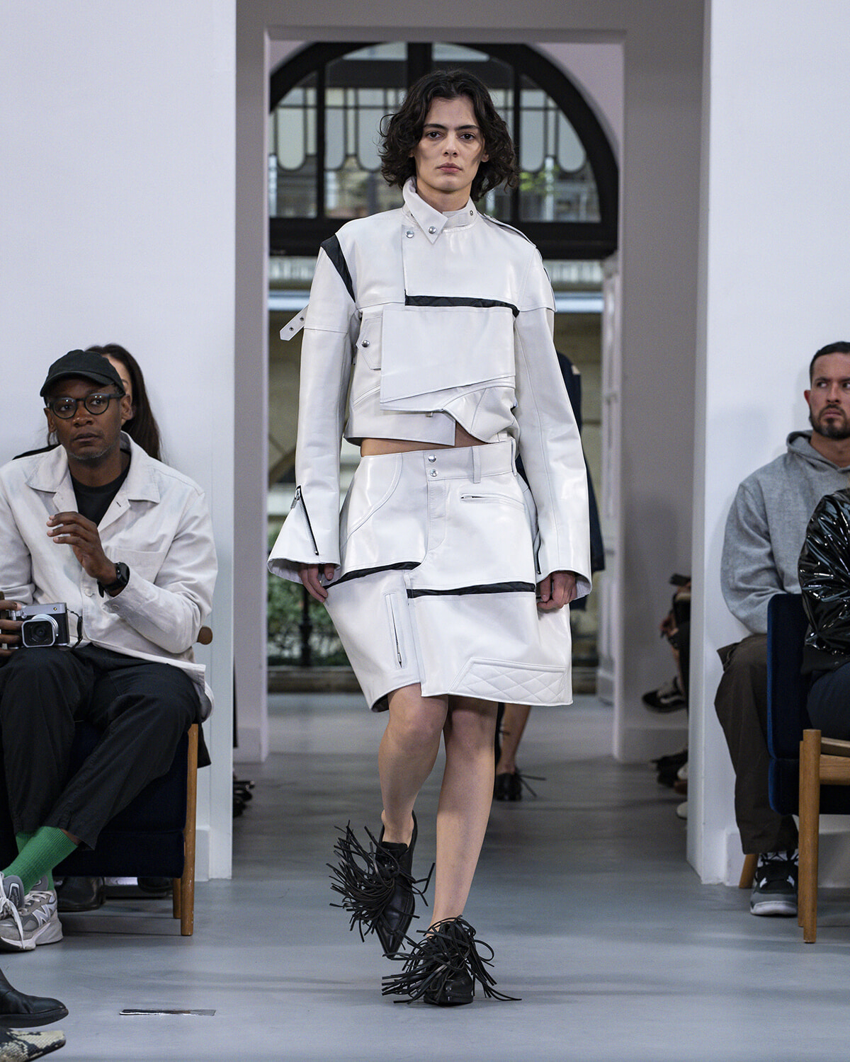 Model walks a white, geometric outfit with black accents on a fashion runway, spectators seated at sides.