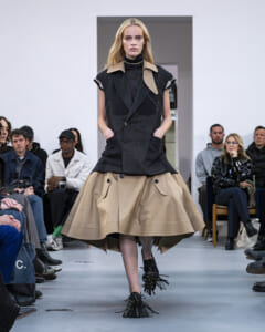 Runway model wearing a black sleeveless double-breasted top with a tan collar and a full beige skirt; audience seated along the runway in the background.