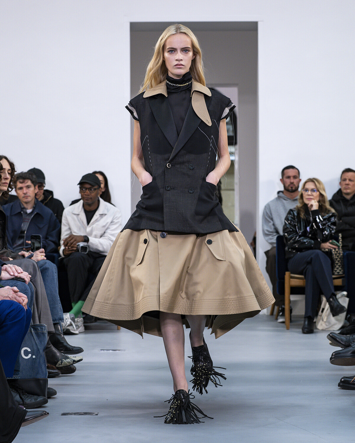 Runway model wearing a black sleeveless double-breasted top with a tan collar and a full beige skirt; audience seated along the runway in the background.
