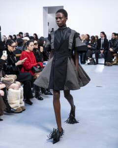 Model walks the runway in a black and gray oversized coat with fringe black shoes; audience seated along the sides.