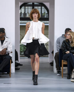 Model walks a fashion runway in a white sleeveless ruffled top and black asymmetric mini skirt with black ankle boots; audience seated along the sides