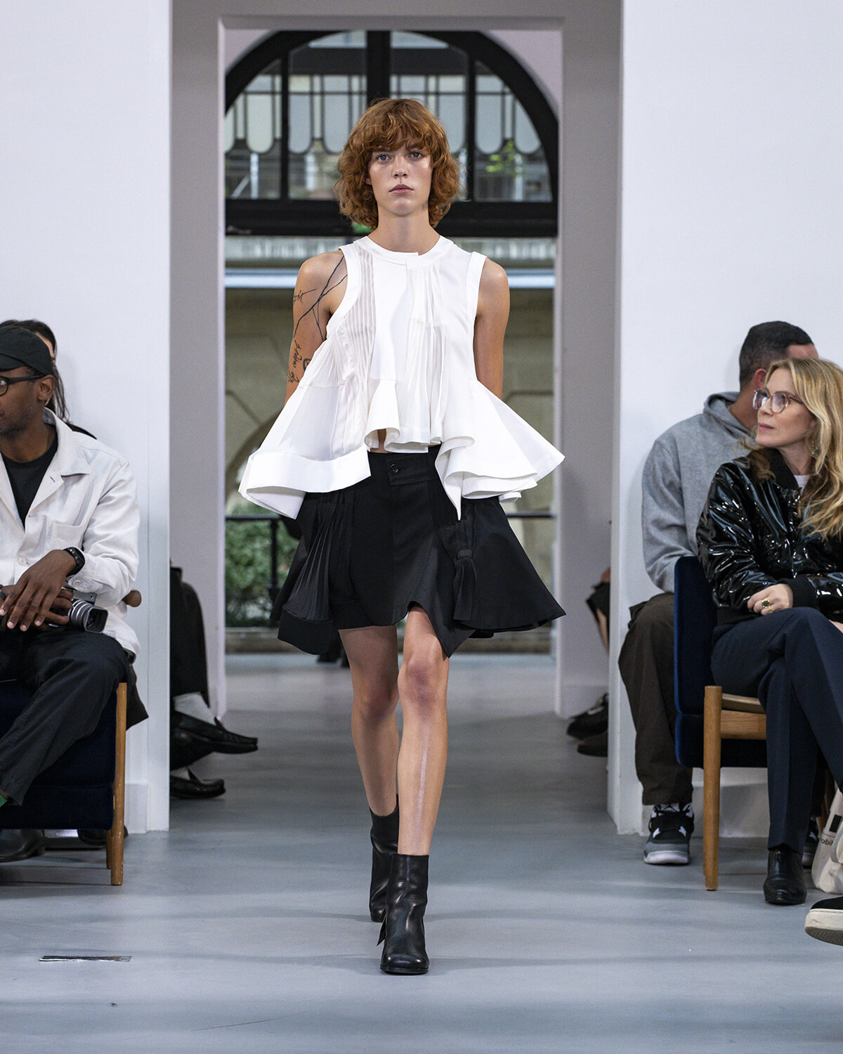 Model walks a fashion runway in a white sleeveless ruffled top and black asymmetric mini skirt with black ankle boots; audience seated along the sides