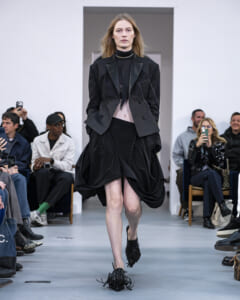 Model walks a fashion runway in a black blazer and draped skirt, audience watching and filming in the background.