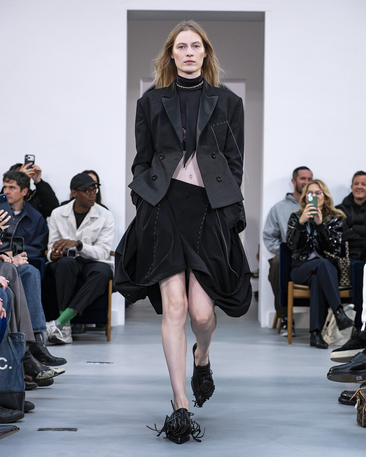Model walks a fashion runway in a black blazer and draped skirt, audience watching and filming in the background.