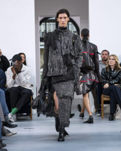 Model walking a runway in a textured gray fringed ensemble, carrying a black handbag and wearing black ankle boots.