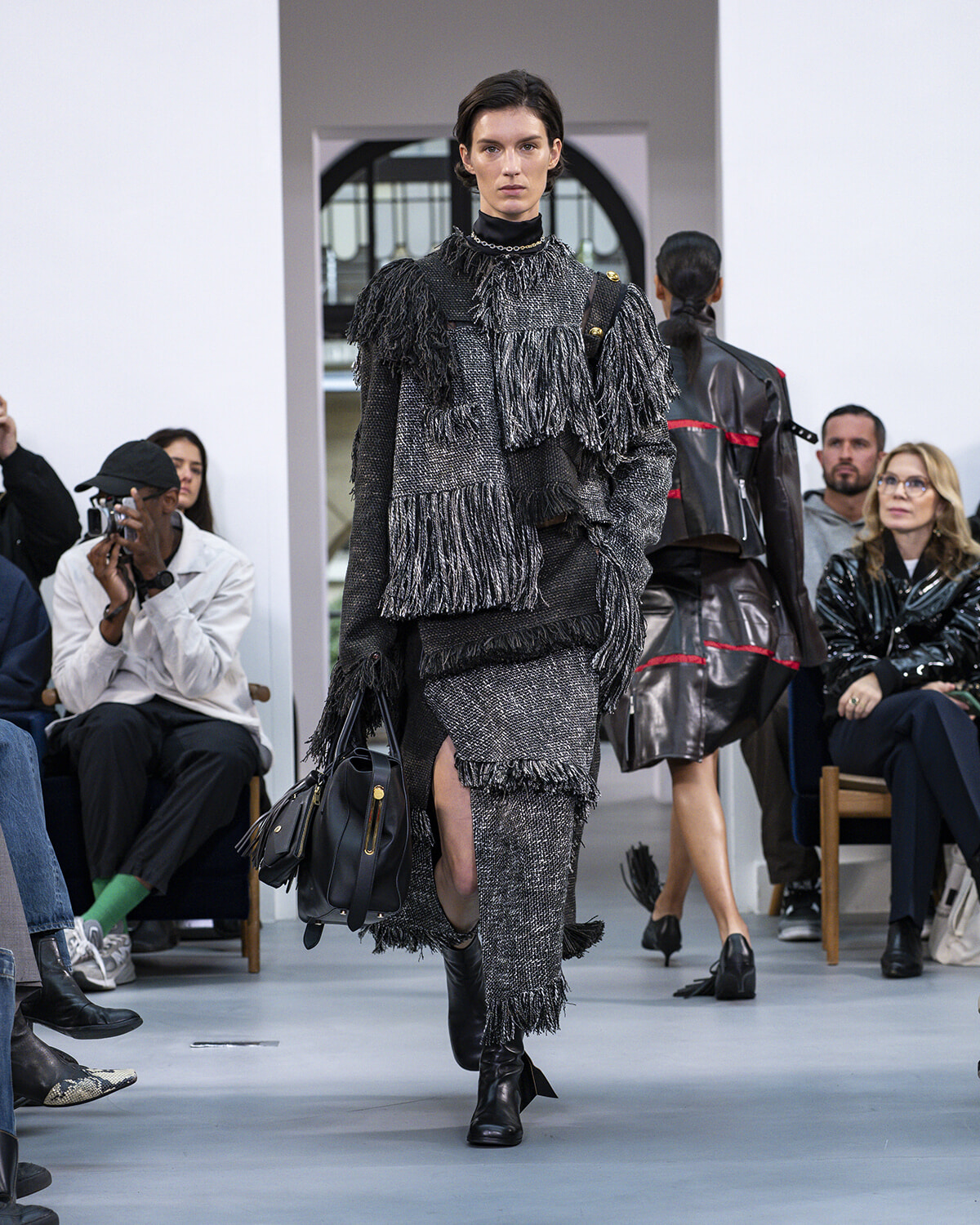 Model walking a runway in a textured gray fringed ensemble, carrying a black handbag and wearing black ankle boots.