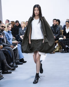 Model walks a fashion show in an oversized olive cape over a white top and green shorts; black ankle boots, seated audience in background.