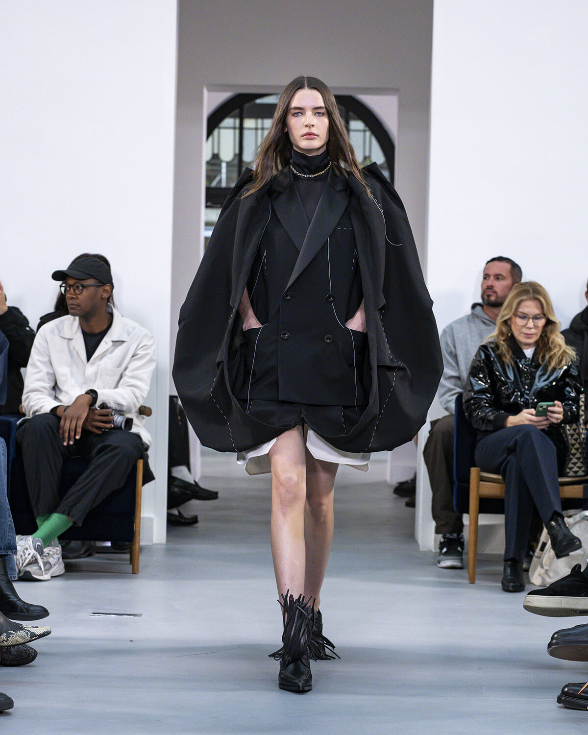 Model walks the runway in an oversized black double-breasted coat with a cape-like silhouette, black turtleneck and short shorts, paired with fringed ankle boots.