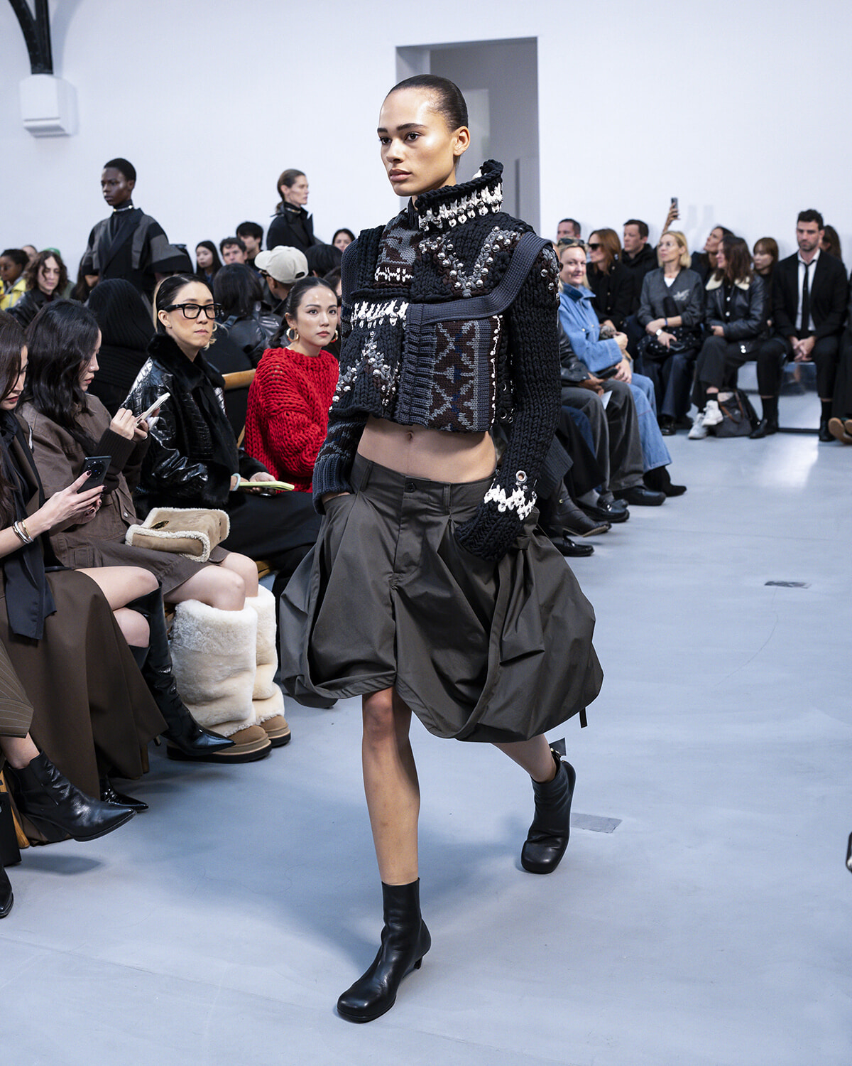 Model walks runway in a textured black knit top with metallic details and a voluminous gray skirt; seated audience watches the show.