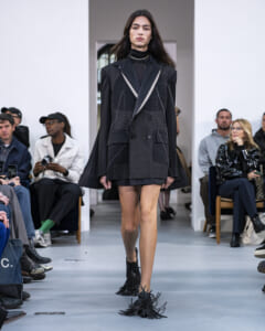 Model walking a fashion show runway in a dark oversized blazer with white stitching, black turtleneck, and fringed ankle boots; seated audience on both sides.