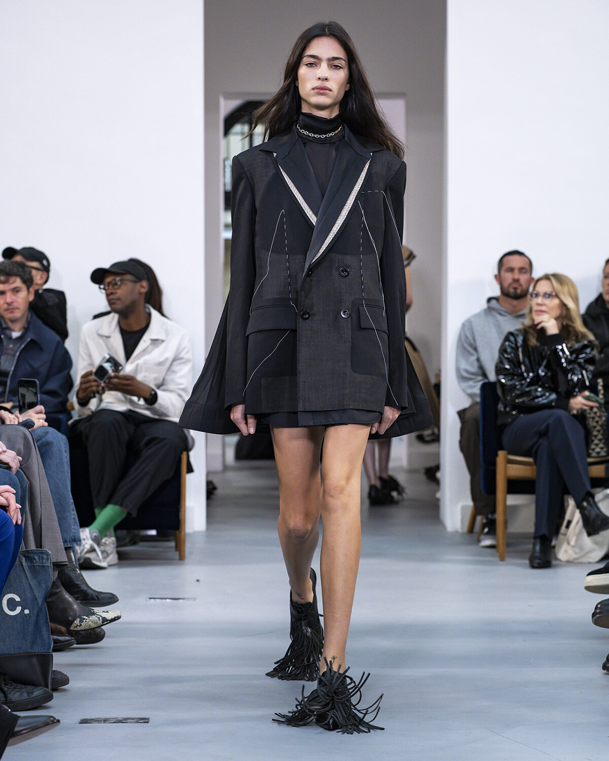 Model walking a fashion show runway in a dark oversized blazer with white stitching, black turtleneck, and fringed ankle boots; seated audience on both sides.