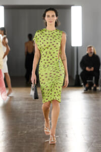 Model walks a runway in a lime-green floral midi dress with ruching, carrying a small black handbag and wearing white strappy sandals.