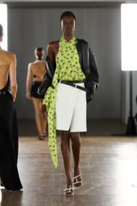 Model walking a runway in a lime green floral wrap top, white shorts, and a black leather jacket draped over the shoulders.