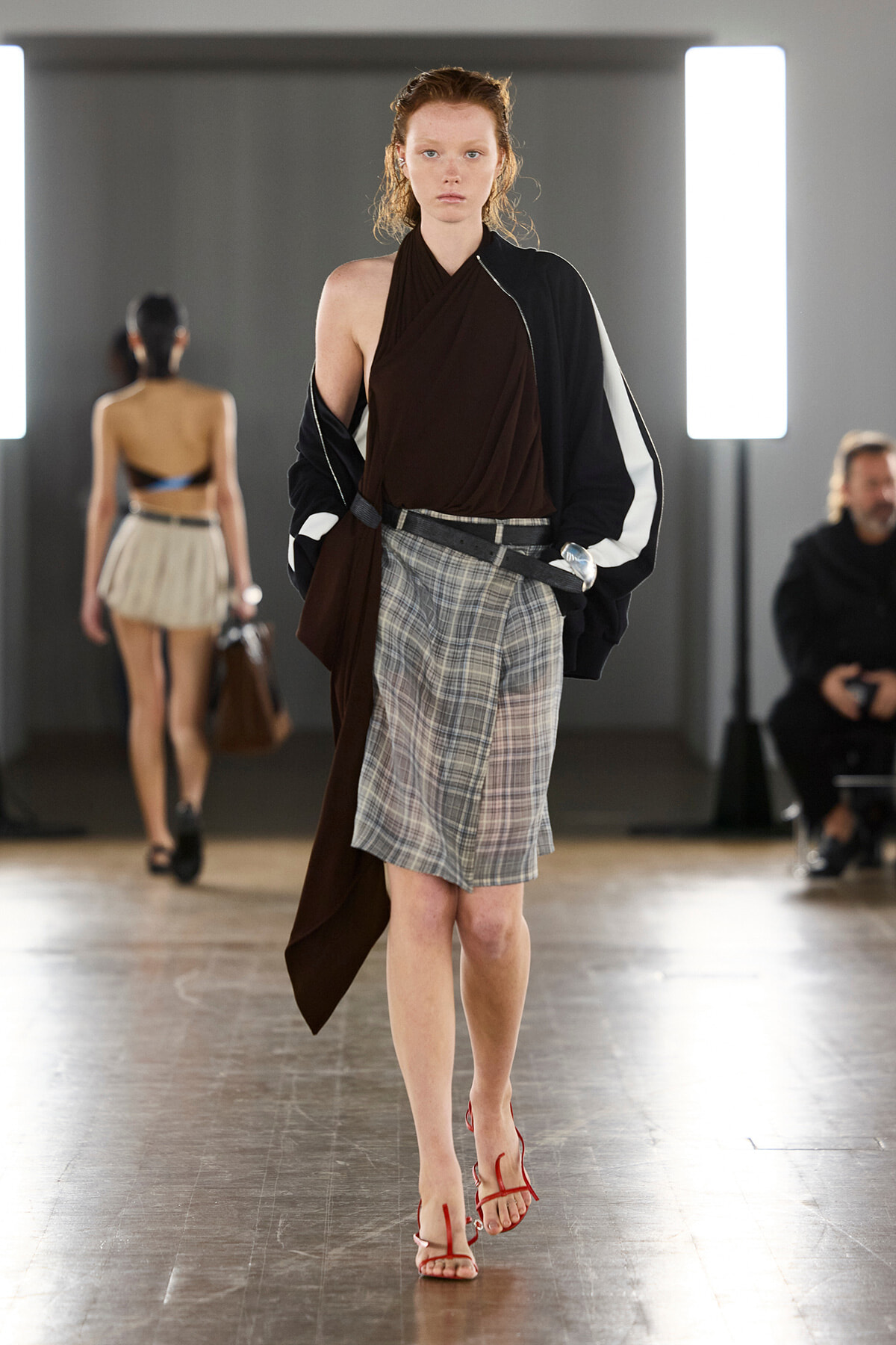 Model walks the runway in a brown draped top, plaid skirt, and a black-and-white jacket, with red strappy sandals.