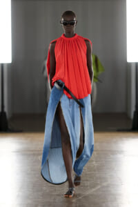 Model walking a runway in a bright red sleeveless pleated top and a long split-denim skirt with a black belt, wearing sunglasses in a studio setting.