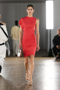 Model walks a red, one-shoulder ruched dress on a runway, carrying a small brown handbag and wearing white strappy heels.