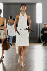 Model walking a fashion runway in a white knit halter top layered over a white knee-length dress, paired with white strappy sandals.