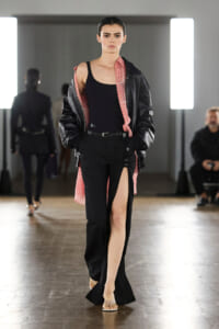 Runway model walking in a black outfit with a high slit, paired with a pink plaid shirt and a black leather jacket used as outerwear.