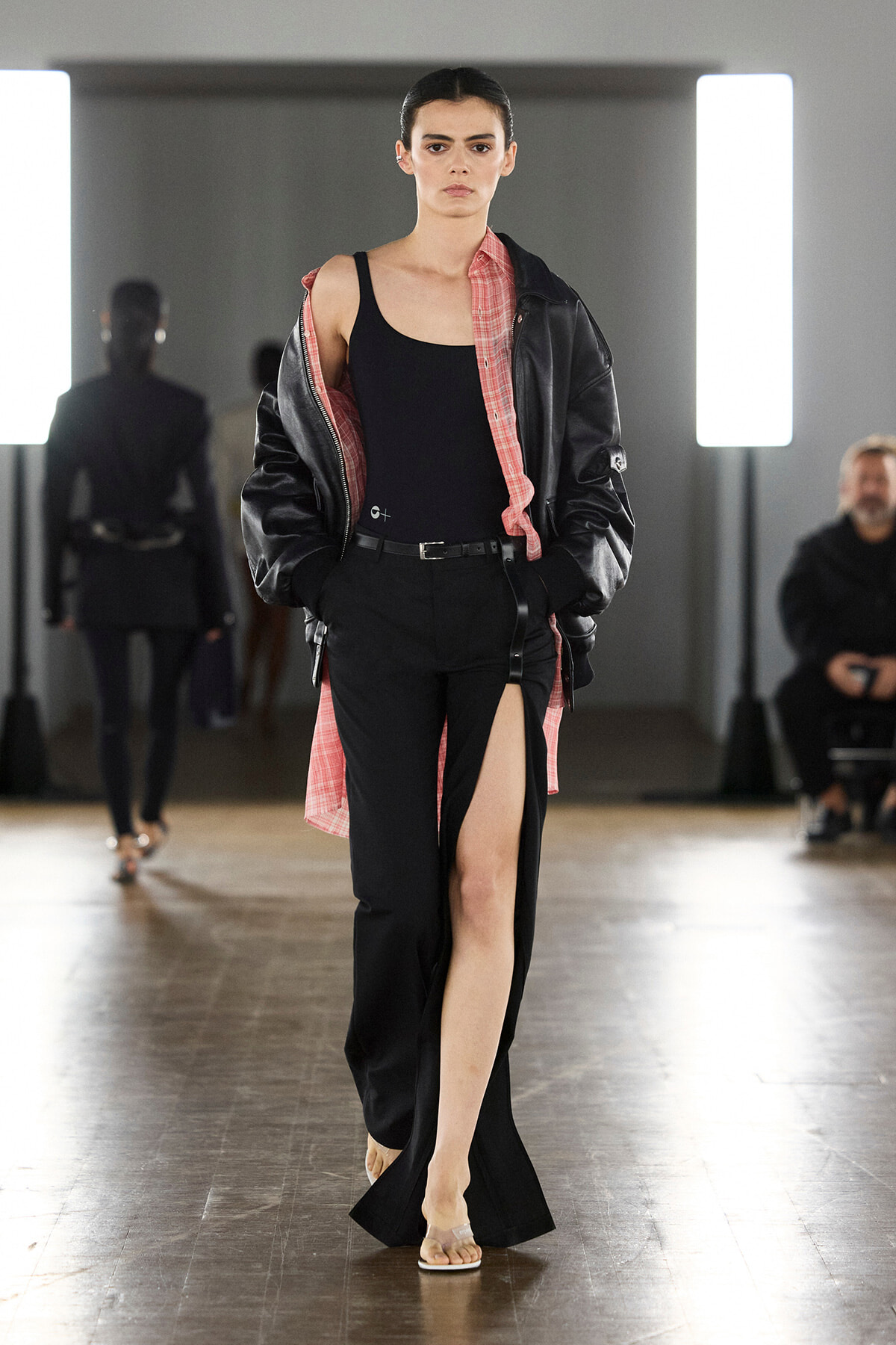 Runway model walking in a black outfit with a high slit, paired with a pink plaid shirt and a black leather jacket used as outerwear.
