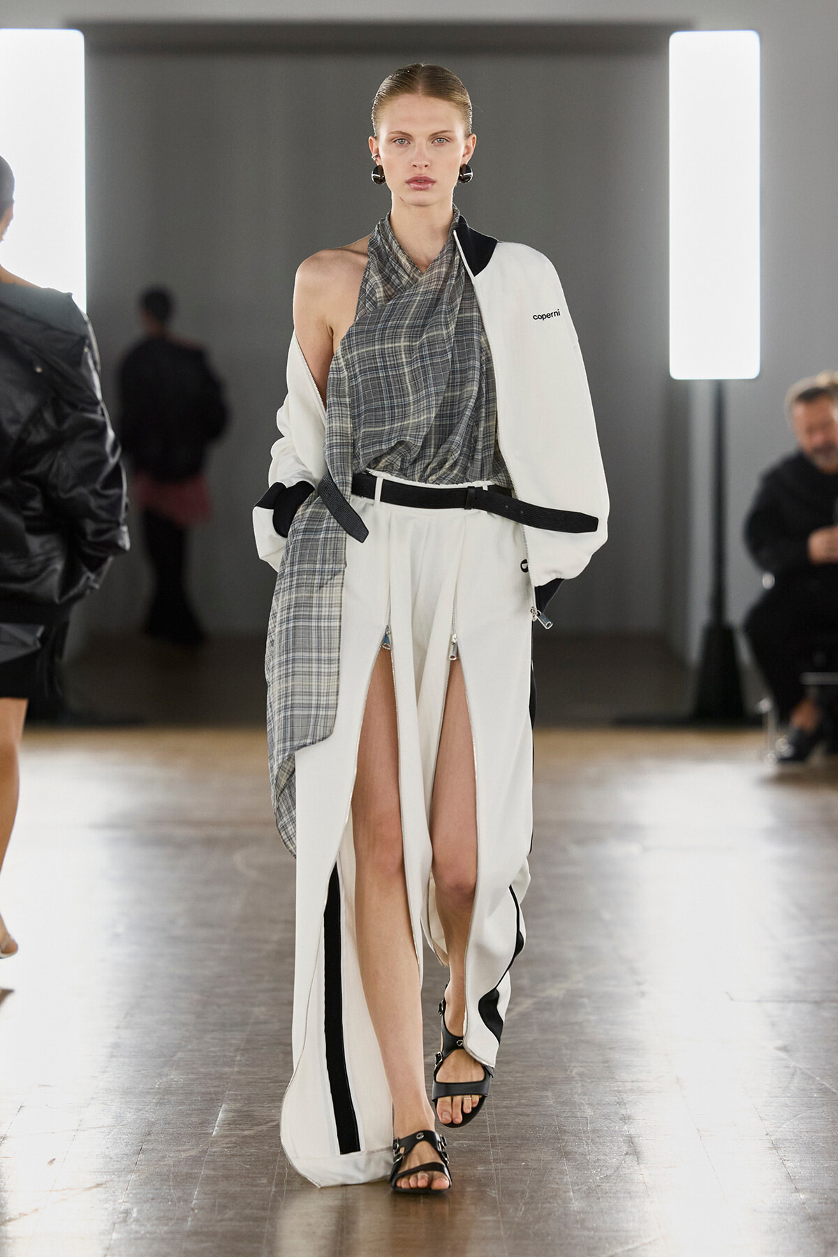 Model walking a fashion runway in a white oversized coat with a gray plaid scarf draped over the front. Reference look includes a black belt and black sandals.