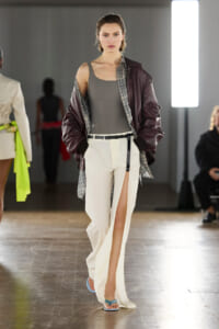 Model walks a runway wearing a gray sleeveless top, oversized burgundy bomber, plaid shirt, and cream wide-leg pants with a front slit and blue sandals.