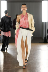 Runway model wearing a pink halter top with a beige jacket draped off one shoulder and white high-slit pants with a black belt and sandals.