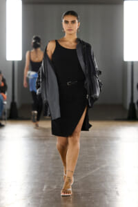 Model wearing a black sleeveless dress with a high slit, gray jacket draped over shoulders, walking a runway with bright studio lights behind.