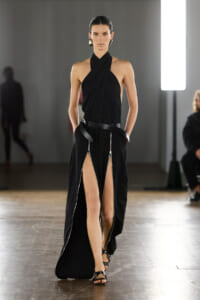 Model walks a runway in a black draped halter gown with high slits and a wide belt, accessorized with bold earrings.