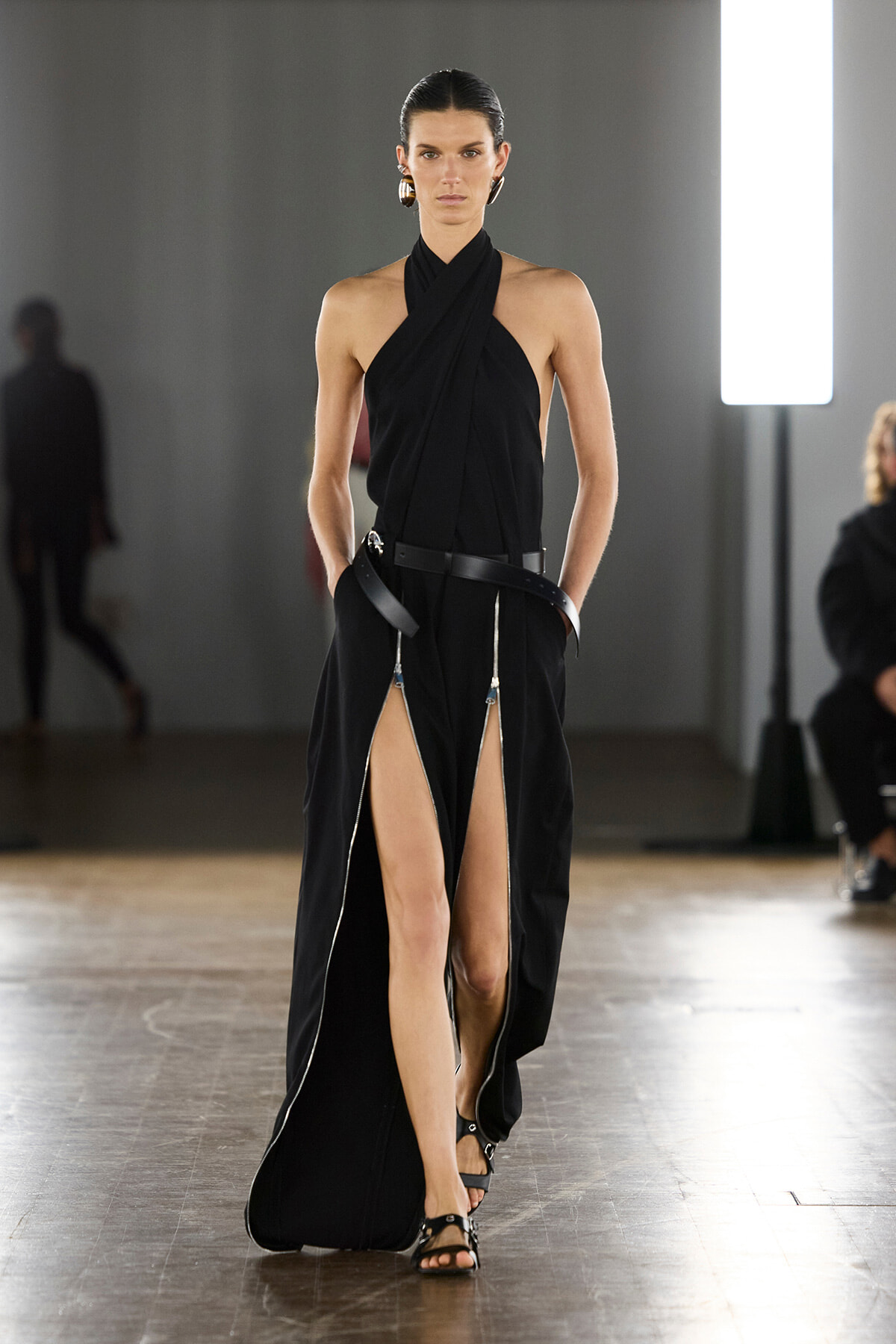 Model walks a runway in a black draped halter gown with high slits and a wide belt, accessorized with bold earrings.