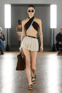 Model on a runway wearing a brown twisted halter top with blue trim and a cream pleated mini skirt, carrying a large brown tote.