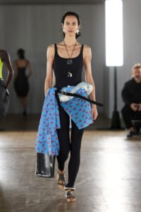 Female model walking a runway in a black sleeveless outfit with a blue floral scarf tied around the waist, black leggings, and black sandals; bold jewelry and a large bag visible to the side.