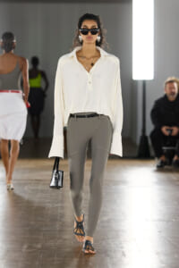 Model walking a fashion runway in a loose white button-up blouse, gray fitted pants, and black sandals, carrying a small black handbag and wearing chunky sunglasses and large silver earrings.