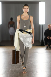 Model walking a fashion runway in a gray bodysuit with a white lace wrap tied at the waist, secured by a black belt, and carrying a tall brown tote.
