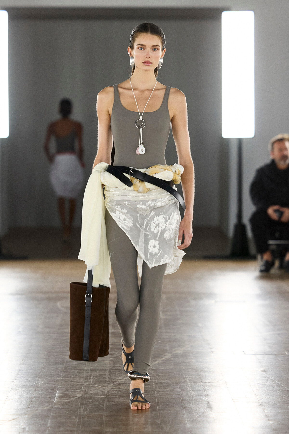 Model walking a fashion runway in a gray bodysuit with a white lace wrap tied at the waist, secured by a black belt, and carrying a tall brown tote.
