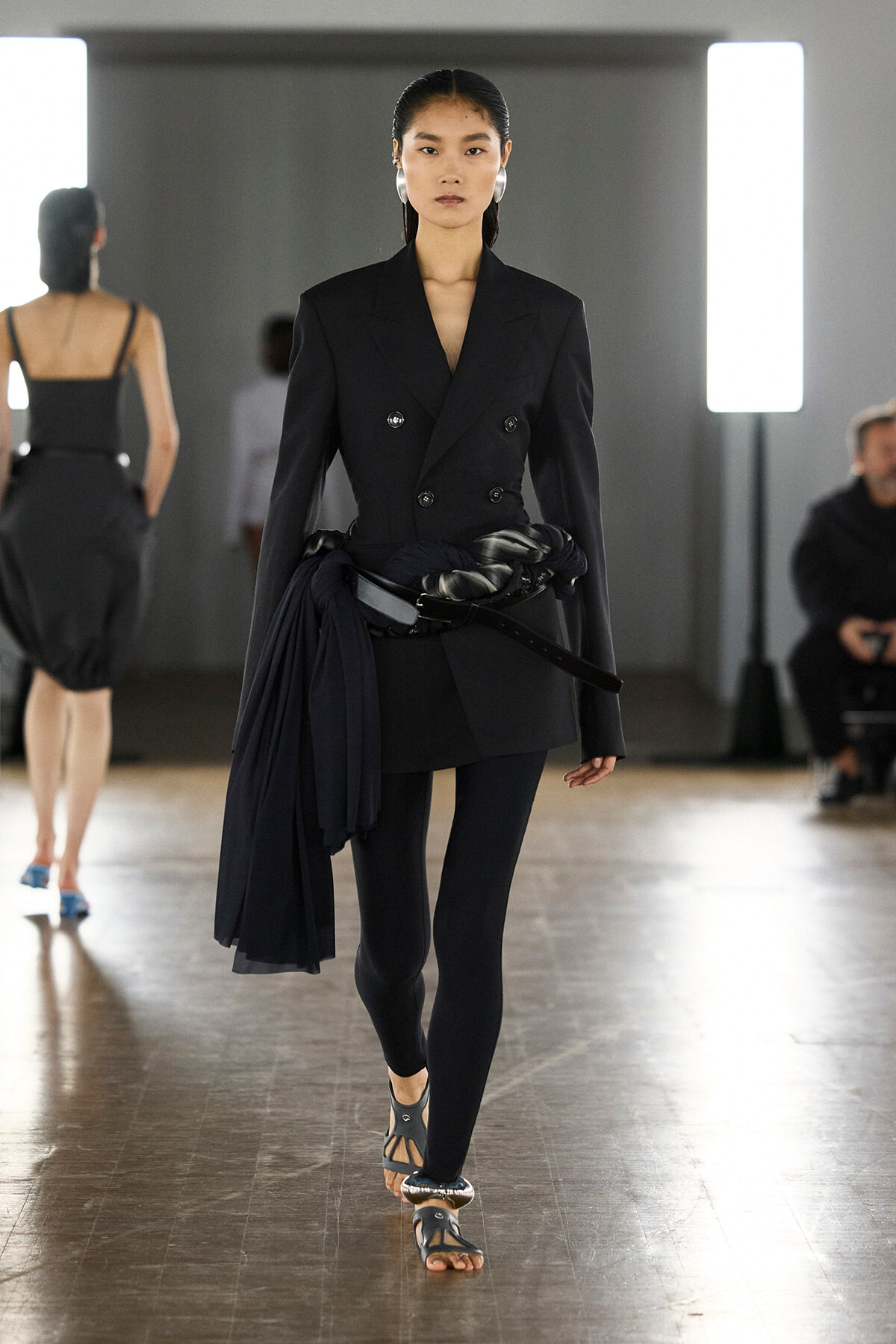Model walking a fashion runway in a dark tailored double-breasted blazer with a fabric belt, black leggings, and sandals in a minimalist backstage setting.