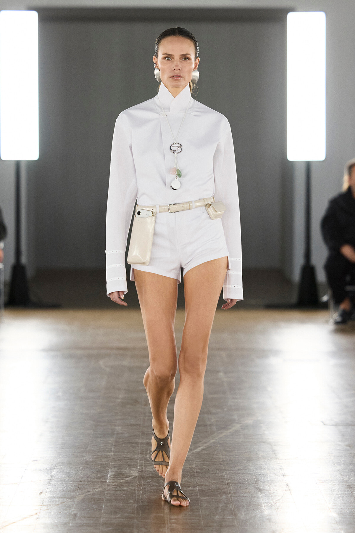 Female model walking a fashion runway in a white high-neck blouse, white shorts, and a beige belt with a pouch, paired with brown sandals.