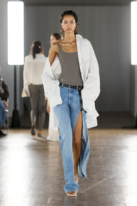 Model walking a runway in a gray tank top, oversized white shirt, and high-slit blue jeans with a black belt and sandals on a wooden floor.