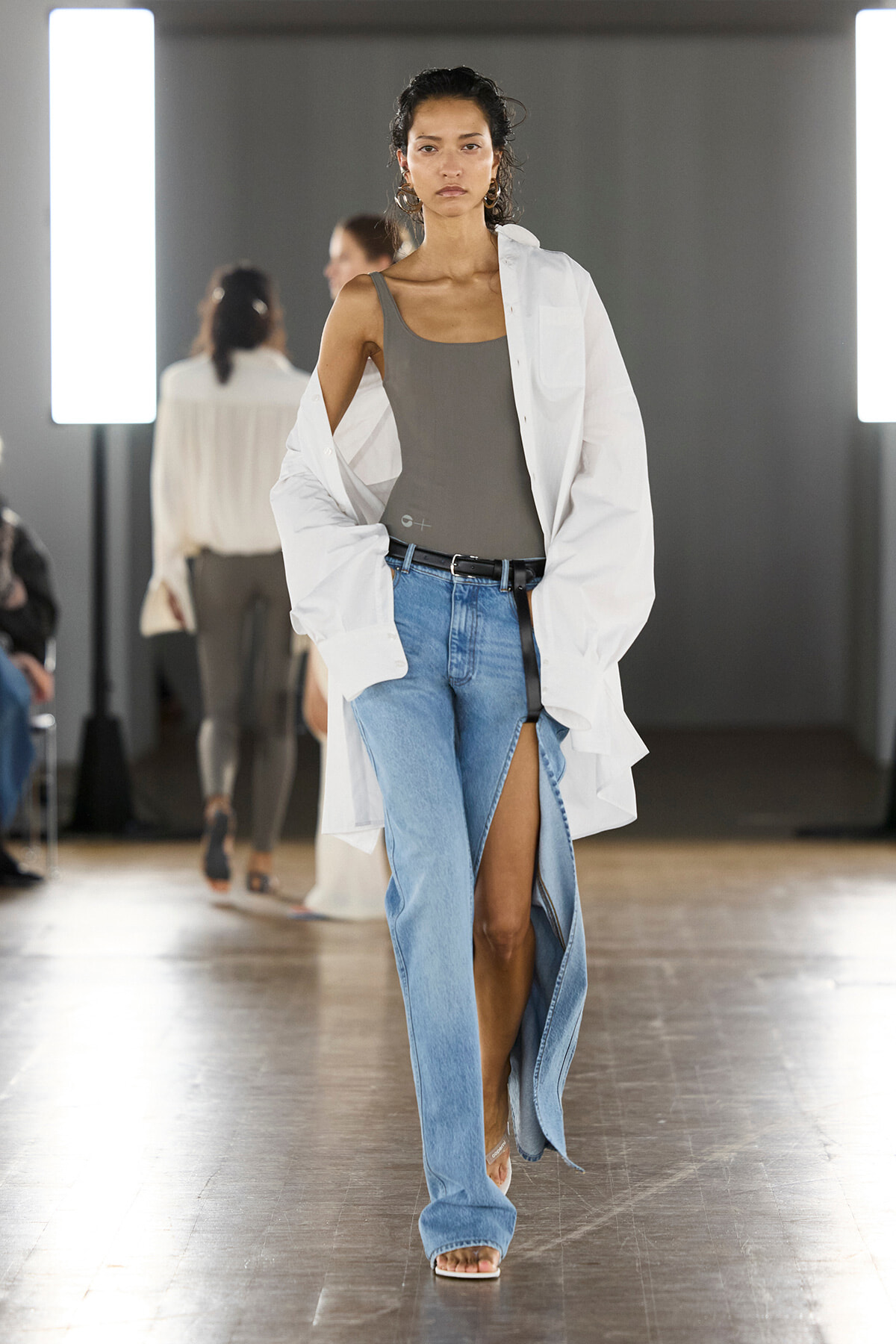 Model walking a runway in a gray tank top, oversized white shirt, and high-slit blue jeans with a black belt and sandals on a wooden floor.