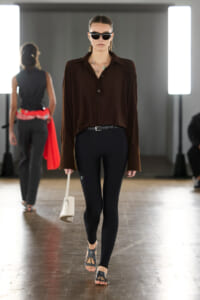 Model walks the runway wearing a brown button-up blouse, black leggings, a black belt, sandals, sunglasses, and a small white handbag on a studio set.
