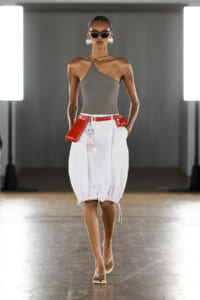 Model walking a runway in a grey asymmetrical halter top, white balloon skirt, and a bold red belt with matching red pouch accessories.