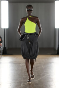 Model walking the runway in a neon lime green one-shoulder top and gray balloon skirt, with a belt bag and sunglasses.