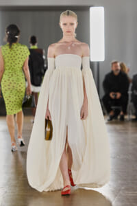 Model on a runway wearing an ivory off-shoulder, floor-length gown with billowing sleeves and a small brown purse; red flat sandals visible beneath the dress.