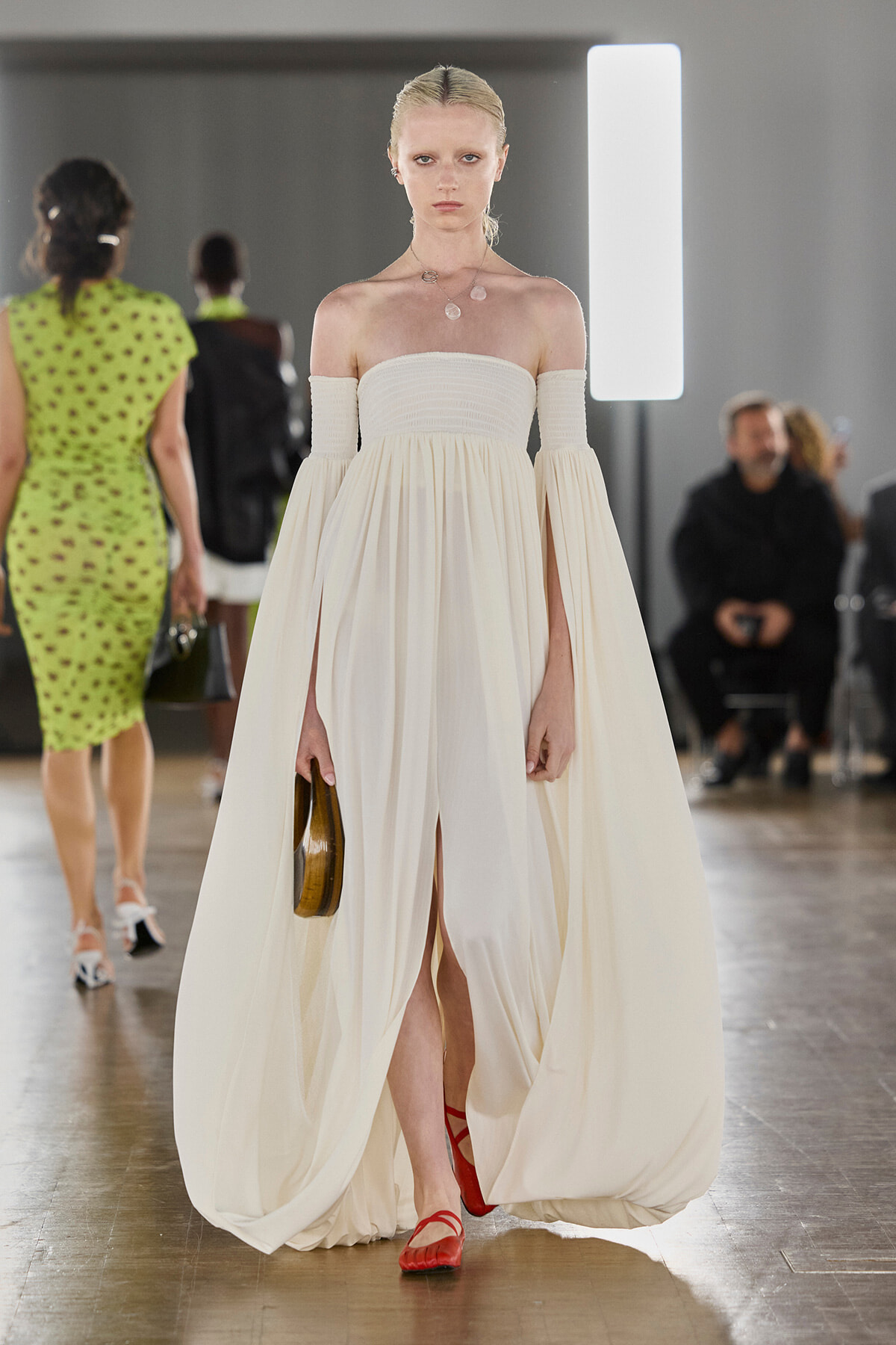 Model on a runway wearing an ivory off-shoulder, floor-length gown with billowing sleeves and a small brown purse; red flat sandals visible beneath the dress.