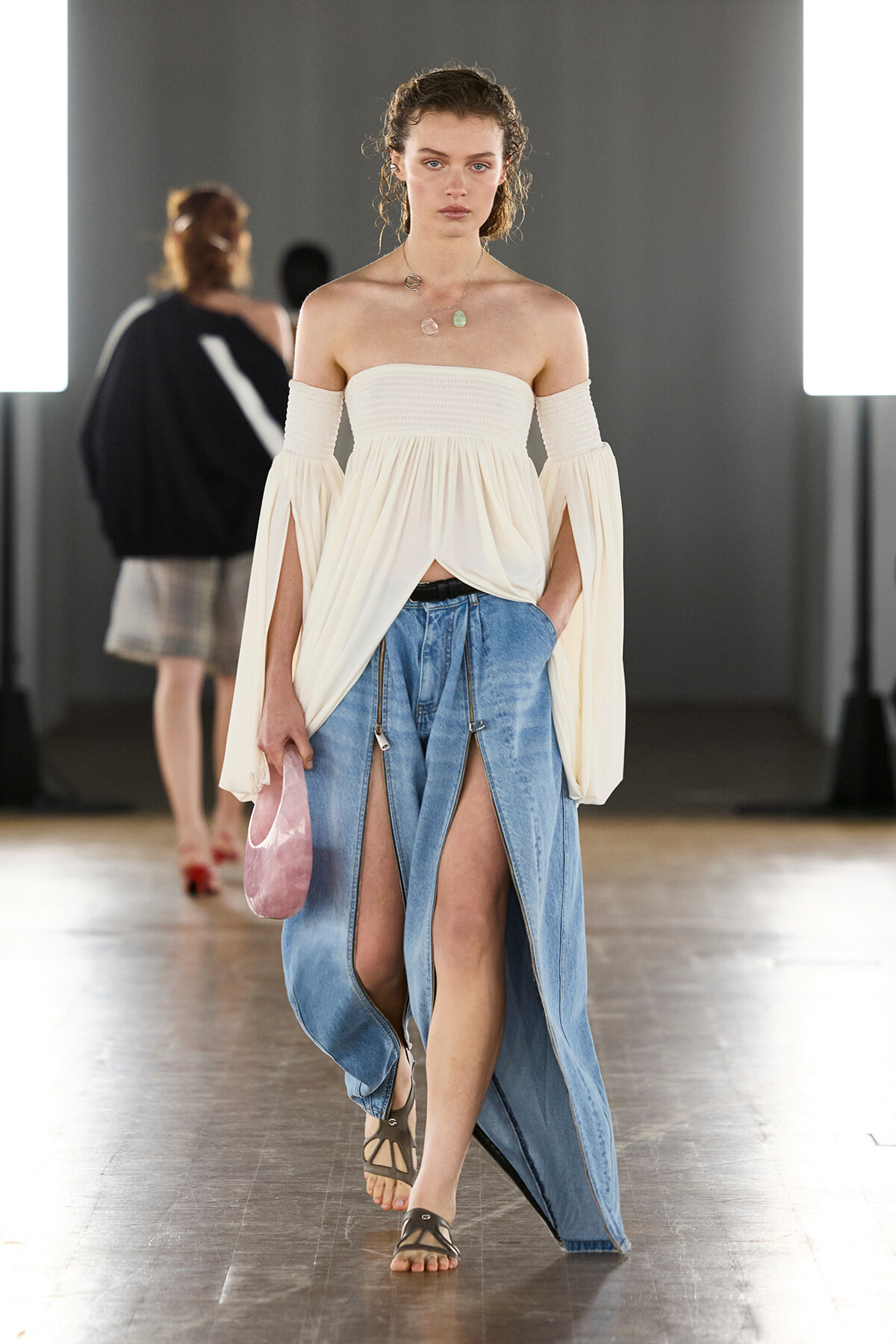 Model walking a runway in an off-shoulder white top with dramatic bell sleeves and a long denim skirt with front slits, holding a small pink bag.