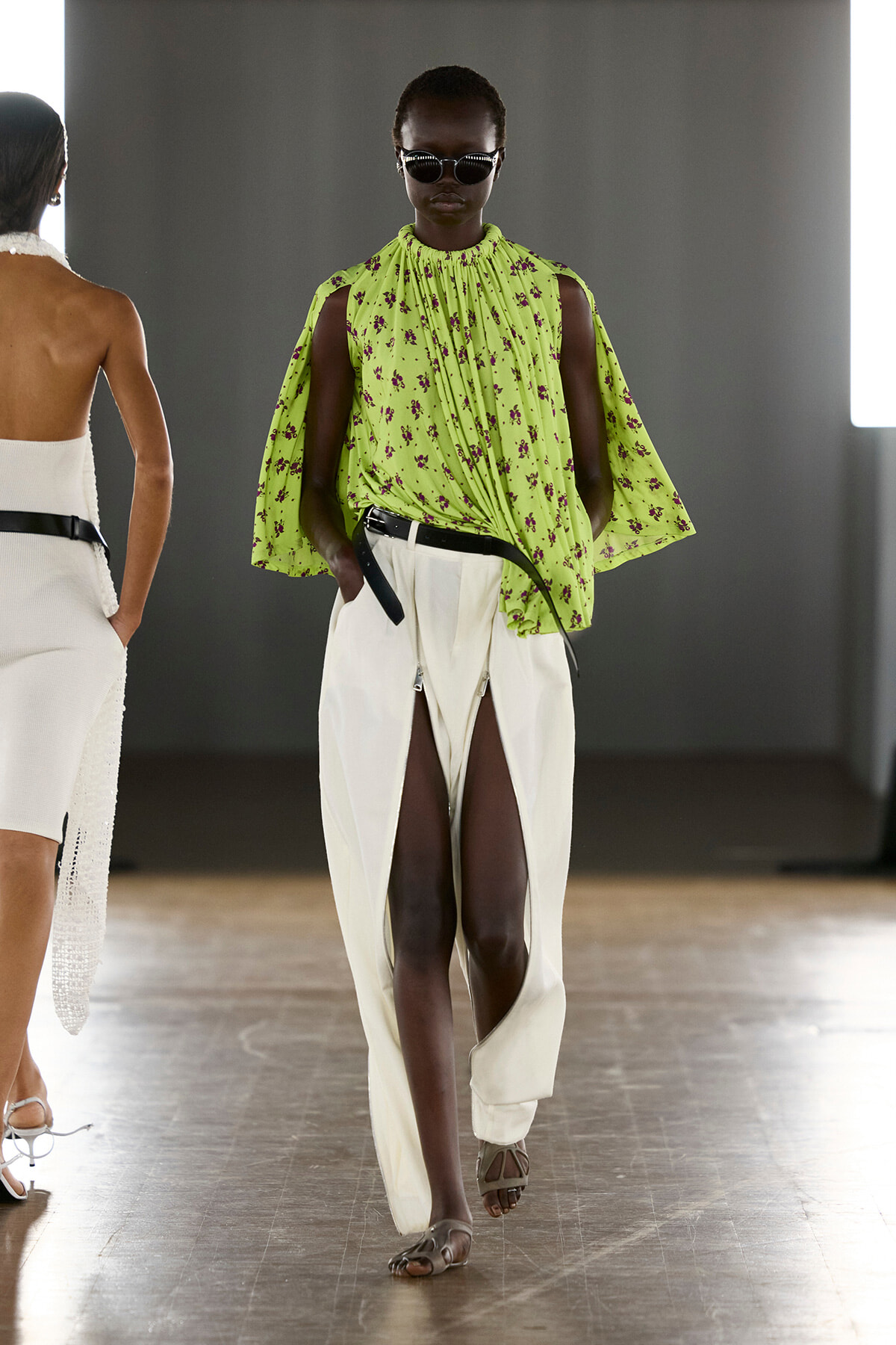 Model on a runway wearing a lime-green floral cape-sleeve top and white split-front pants with sandals, sunglasses.
